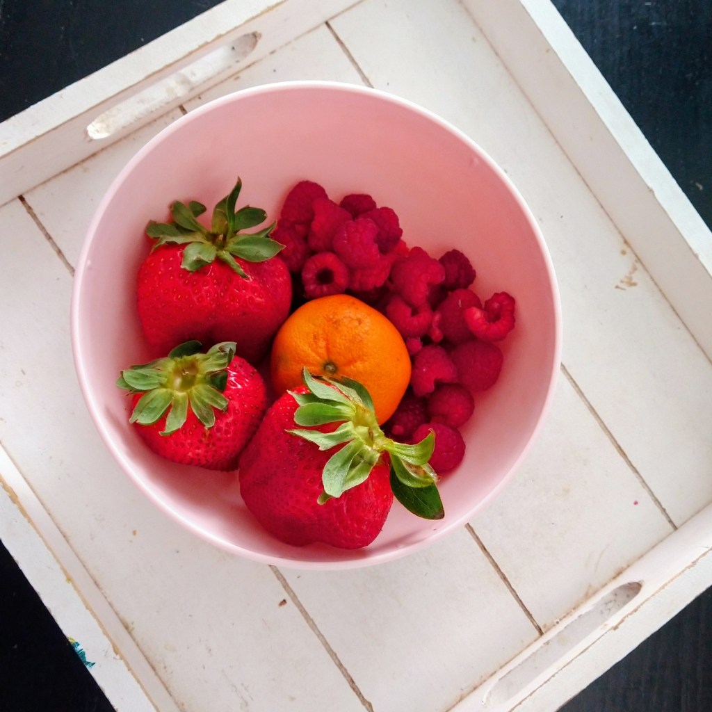 Red raspberries and strawberries with a clementine orange in the middle of a pink bowl on a weathered white wood tray.